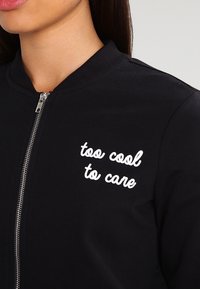 Close-up van een persoon in een zwarte zip-up jas met op de borst witte geborduurde tekst die zegt "te cool om je druk te maken".