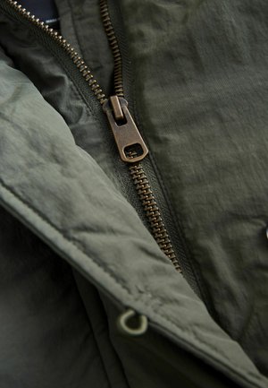 Olijfgroene jas met een opvallende bronzen ritssluiting, een gestructureerde stof en gestikte naaddetails langs de randen.