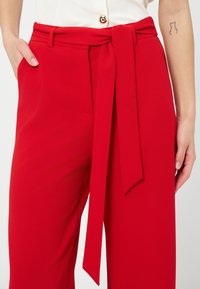 Femme portant un pantalon large rouge taille haute avec une ceinture en tissu nouée, un haut blanc boutonné, et un petit tatouage en forme de contour de chat sur l'avant-bras gauche.
