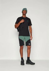 Vīrietis, kurš valkā zaļu cepuri, melnu t-kreklu, zaļus un melnus šortus, melnas zeķes un apavus, tur melnas mugursomas siksnu, stāv uz pelēka grīdas.