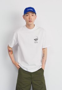 Hvid kortærmet t-shirt med grafisk print på venstre bryst. Olivengrønne shorts. Blå kasket med tekst, båret fremad. Minimalistisk design.