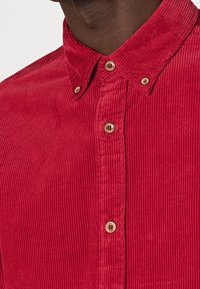 Camisa de pana roja con cuello abotonado, que presenta una textura de canalé vertical y botones dorados a lo largo de la parte delantera.