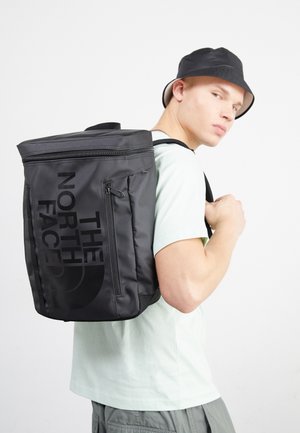 Jeune homme portant un chapeau bob noir, avec un grand sac à dos noir The North Face porté sur une épaule, vêtu d'une chemise claire et d'un pantalon gris.