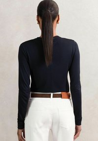 Donna con capelli scuri e lisci raccolti in una coda di cavallo, indossa un top nero a maniche lunghe infilato in pantaloni bianchi con una cintura marrone, in piedi davanti a uno sfondo semplice.