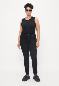 Black sleeveless top paired with black slim-fit jeans. Model wears chunky black shoes and sunglasses, showcasing a sleek, monochrome look.