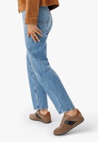 Brązowe zamszowe sneakersy z czarnymi akcentami, w połączeniu z jasnoniebieskimi, luźnymi dżinsami z surowym dołem oraz brązową zamszową kurtką.