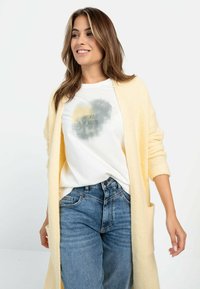 Lys gul strikket cardigan med lange ærmer, båret over en hvid t-shirt med et gråt og gult grafik, matchet med blå denimjeans.