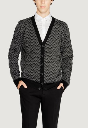 Cardigan a motivi chevron bianco e nero con scollo a V, chiusura con bottoni e polsini a costine, indossato sopra una camicia bianca.