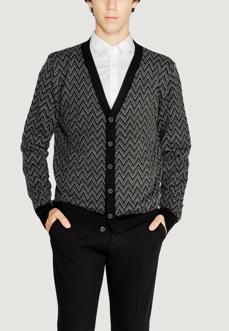 Cardigan a motivi chevron bianco e nero con scollo a V, chiusura con bottoni e polsini a costine, indossato sopra una camicia bianca.