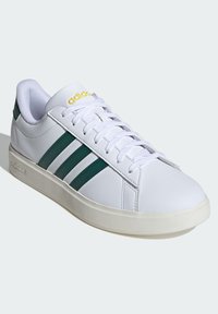 Biele tenisky so zelenými pruhmi a žltým logom. Má hladký kožený vrch, šnurovanie, polstrovaný golier a textúrovanú gumovú podrážku.