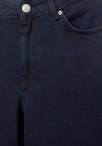 Gros plan sur un jean en denim bleu foncé montrant le bouton avant, la braguette à fermeture éclair, les passants de ceinture et les détails de couture des poches.