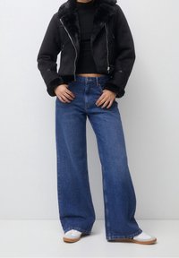 Personne portant une veste courte en fausse fourrure noire, un haut côtelé noir, un jean bleu à jambes larges et des baskets blanches sur un fond uni.