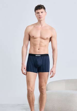 Shirtless man standing, wearing navy blue boxer briefs with white vertical stripes and black waistband labeled "BOSS," beside a white textured chair.