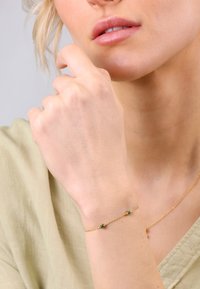 Goldarmband mit grünen Edelsteinen an einer filigranen Kette, getragen an einer Hand mit feiner Hautstruktur und neutraler Kleidung.