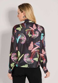Frau mit blonden Haaren, die eine schwarze Satinbluse mit buntem Schmetterlingsdruck trägt, von hinten gesehen, kombiniert mit schwarzen Hosen.