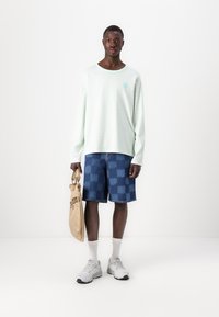 Light mint green long-sleeve shirt with stripes, paired with blue patterned denim shorts. Holding a beige bag with a strap, wearing white sneakers.