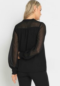 Blusa negra con mangas transparentes de puntos, detalle fruncido en la parte trasera y puños elásticos. Textura suave con un ajuste relajado.