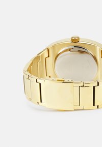 Orologio da polso dorato con bracciale a maglie metalliche, cassa rotonda, finitura texturizzata e vetro rotondo. Include una corona per la regolazione.