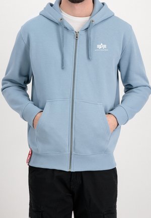 Mężczyzna ubrany w jasnoniebieską bluzę z kapturem na zamek błyskawiczny z przednimi kieszeniami i logo "Alpha Industries" na piersi, w połączeniu z czarnymi spodniami.