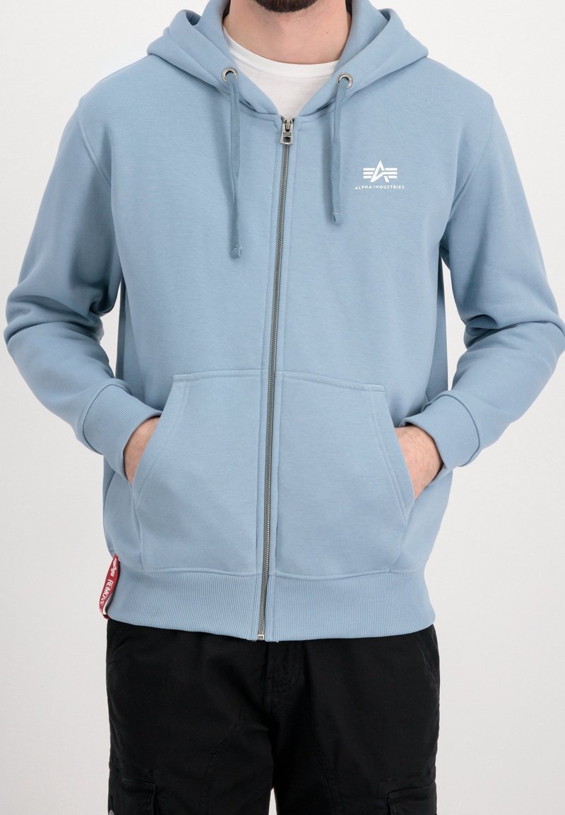 Homme portant un sweat à capuche bleu clair zippé avec des poches avant et le logo "Alpha Industries" sur la poitrine, assorti à un pantalon noir.