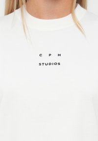 Hvit bomull T-skjorte med svart tekst som leser "C P H" over "STUDIOS" i en enkel, moderne skrifttype. Myk tekstur og rund hals.