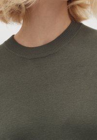 Olivgrön tröja med ribbat rundad halsringning och texturerad tyg, med fokus på en nära bild av kragen och övre bröstområdet.