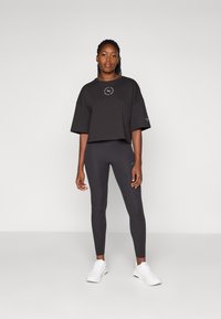 Černé zkrácené tričko s kruhovým logem na hrudi, doplněné tmavými legínami a bílými sportovními botami. Hladká textura a minimalistický design.