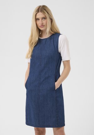 Femme blonde portant une robe en denim bleu sans manches par-dessus une chemise blanche à manches courtes, debout, les mains dans les poches de la robe, sur un fond uni.