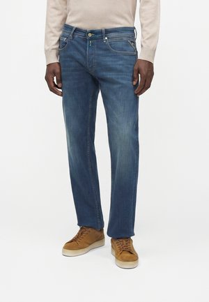 Person wearing blue jeans, beige long-sleeve top, and brown suede sneakers, standing against a plain white background.