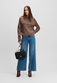 Brauner, gerippter Pullover mit "Karl"-Stickerei, kombiniert mit weit geschnittenen blauen Jeans. Dazu eine schwarze, gep quiltete Handtasche und dunkle, hochhackige Stiefel.