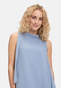 Lichtblauw mouwloos topje met een gedrapeerd ontwerp, voorzien van een hoge halslijn en zachte, vloeiende stof die de vloeibare uitstraling versterkt.