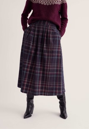 Persona con falda midi de cuadros oscura, jersey de punto granate con patrón y botas negras de tacón, de pie con las manos en los bolsillos.