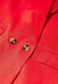 Gros plan d’un blazer rouge vif avec deux boutons ronds en écaille et une poche à rabat sur le tissu.