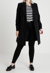 Svart dubbelknäppt kappa med en strukturerad design, bärs över en svart och vit randig topp, parat med svarta skinny byxor och loafers.