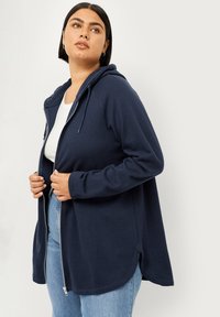 Marineblauwe rits hoodie met een capuchon, lange mouwen en een gebogen zoom. Gemaakt van zachte stof, met een relaxte pasvorm en zijsplitsingen.