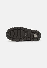 Suole di scarpe in gomma nera con un motivo testurizzato e robusto per una buona aderenza, caratterizzate da un logo circolare e dal nome del marchio su un lato.