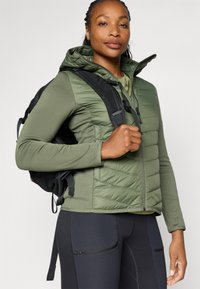 Giacchetto verde isolato con un design trapuntato, caratterizzato da uno stile aderente e un cappuccio, abbinato a leggings neri e uno zaino nero.
