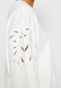 Bílá mikina s jemn�ě vyřezávanými květinovými vzory na rukávech, která má měkkou, hladkou texturu a volný střih.