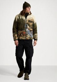 Fleecinė striukė su žaliame, smėlio spalvos ir mėlyname fone vaizduojamu tekstūruotu gamtos peizažu; užsegama užtrauktuku ir turi krūtinės kišenę. Dėvima su juodomis krovinio kelnėmis.