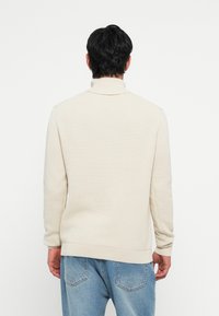 Beige gebreide trui met hoge kraag en ribgeborduurde zoom, gecombineerd met lichtblauwe jeans. Het ontwerp heeft een naadloze achterkant.