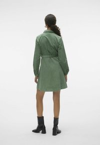 Žena s kudrnatými vlasy v nízkém culíku, oblečená v zelených kordových šatech s páskem a černých kotníkových botách, stojící zády k fotoaparátu na bílém pozadí.