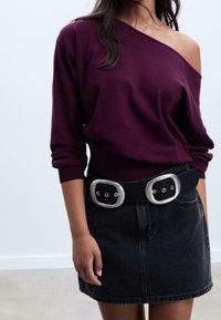 Pull bordeaux à épaules dénudées associé à une jupe en jean noire, comprenant une ceinture large noire avec des boucles et œillets argentés.