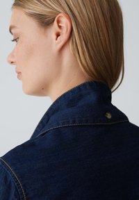 Giacca di jeans con una tonalità blu scuro, dotata di un colletto strutturato, cuciture in vista e una chiusura a un solo bottone al collo.