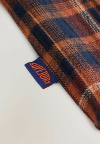 Rutet tekstil i orange, marineblå og beige med en tekstureret overflade. Har et blåt mærke med teksten "Superdry."