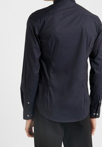 Person som bär en figurnära mörk marinblå skjorta med lång ärm och svarta byxor, sedd bakifrån mot en enkel vit bakgrund.