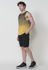 Top z gradientom rumene in sivo z logotipom, v kombinaciji s črnimi kratkimi hlačami s številnimi žepi in črnimi supergami. Gladka tekstura, sproščen dizajn.