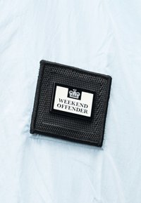 Μαύρο υφασμάτινο ένθετο σε σχήμα τετραγώνου με λευκή ετικέτα και εικόνα κορώνας, το κείμενο γράφει "WEEKEND OFFENDER" σε φόντο ανοιχτού μπλε υφάσματος με ζάρες.