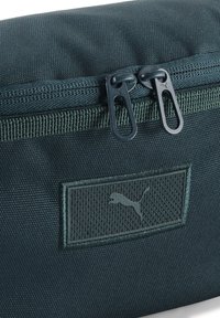 Borsetta in tessuto verde scuro con chiusura a zip, caratterizzata da un pannello testurizzato e un'etichetta con logo a rilievo. Materiale liscio e resistente con doppia cerniera.