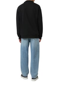 Zwarte sweatshirt met lange mouwen, losse pasvorm. Gecombineerd met lichtblauwe baggy jeans en grijze sneakers, staande van achteren.