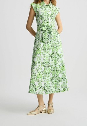 Ärmelloses Midikleid mit Kragen und kompliziertem grün-weißem Blumenmuster, taillierter Gürtel, getragen mit goldenen spitzen Ballerinas mit Knöchelriemen.